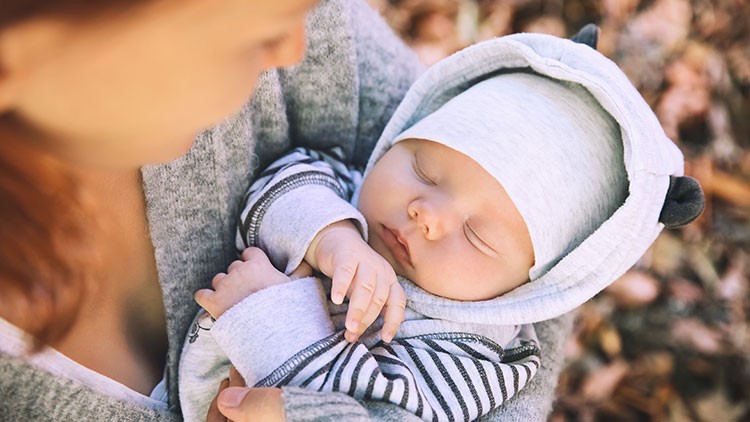 Person in grauer Kleidung hält ein Baby in einem gestreiften und grauen Outfit mit Kapuze, im Hintergrund Herbstlaub.