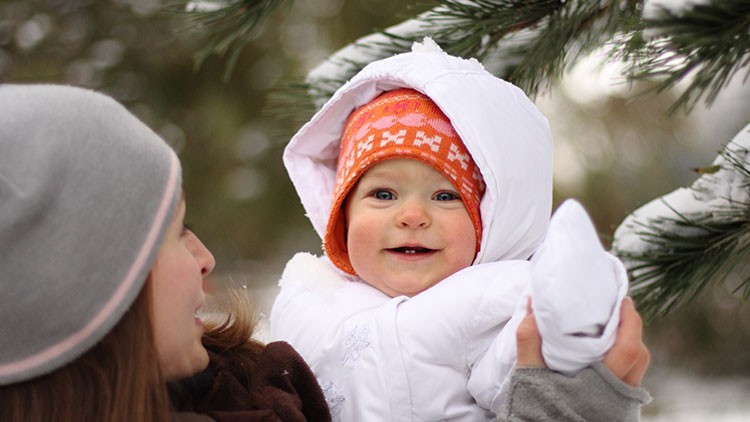 Person hält ein Kind in weißer Winterkleidung und orangefarbener Mütze unter verschneiten Tannenzweigen.