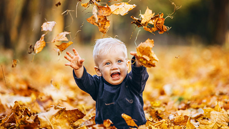 Kind in dunkler Kleidung spielt im herbstlich mit Laub bedeckten Park und wirft Blätter in die Luft.