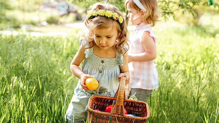 Kind in heller Kleidung und Blumenkranz hält einen Korb mit Obst in einer grünen Wiese, ein weiteres Kind steht dahinter.
