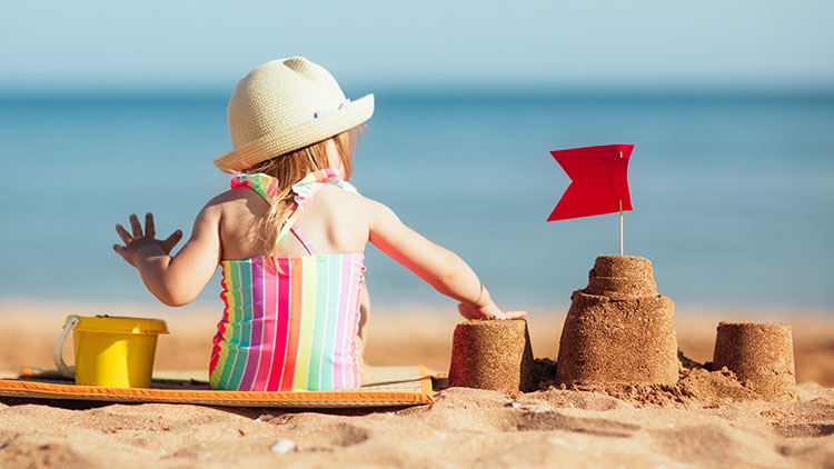 Kind in gestreiftem Badeanzug und Strohhut sitzt am Strand vor einer Sandburg mit roter Fahne, gelber Eimer daneben.