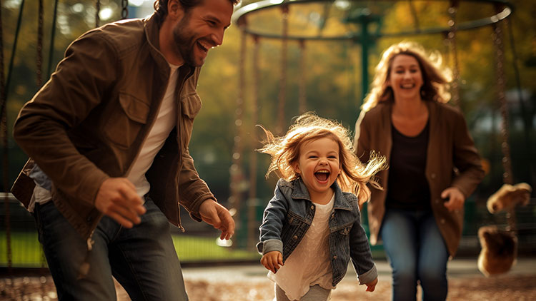 Ein Kind rennt mit zwei Erwachsenen auf einem Spielplatz mit herbstlichem Laub und einer Schaukel im Hintergrund.