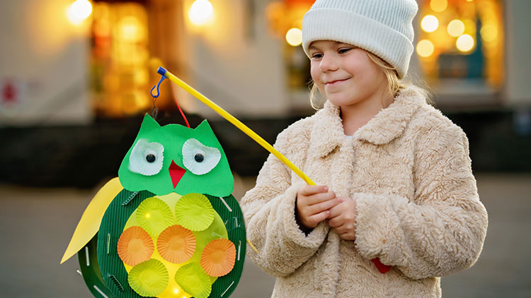 Ein Kind in beiger Jacke hält eine bunte Laterne in Form einer grünen Eule mit gelbem Stab.