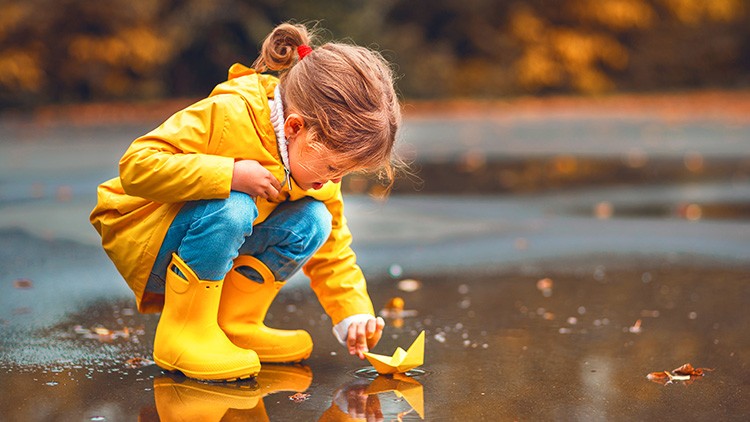 Kind in gelber Regenkleidung kniet an einer Pfütze und lässt ein kleines gelbes Papierboot im Wasser schwimmen.