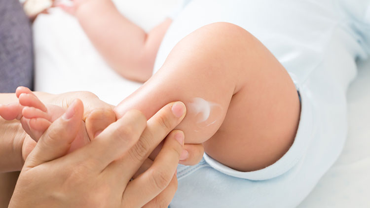Eine Hand trägt Creme auf die Haut eines Babys auf, das helle Kleidung trägt und auf einer weißen Unterlage liegt.