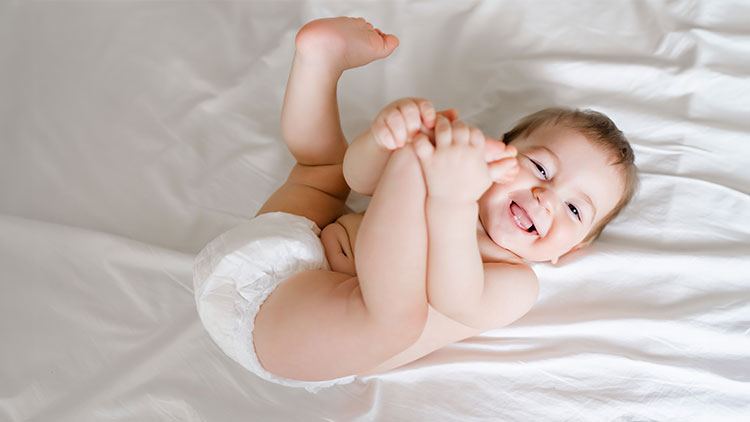 Ein Baby liegt in Windel auf einem weißen Laken und hält mit beiden Händen seine Füße.