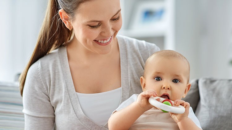 Eine Person in hellem Oberteil hält ein Baby, das etwas Weißes in den Händen hält, auf einem Sofa in einem Wohnzimmer.