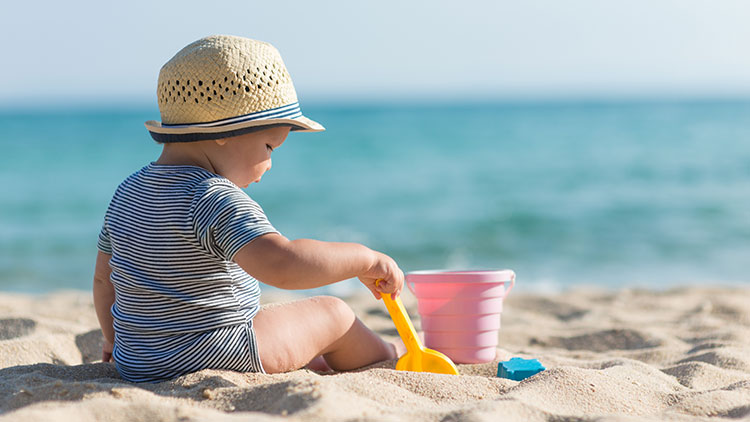 Kleinkind mit Strohhut spielt mit Schaufel, rosa Eimer und Förmchen am Sandstrand, im Hintergrund blauer Ozean.