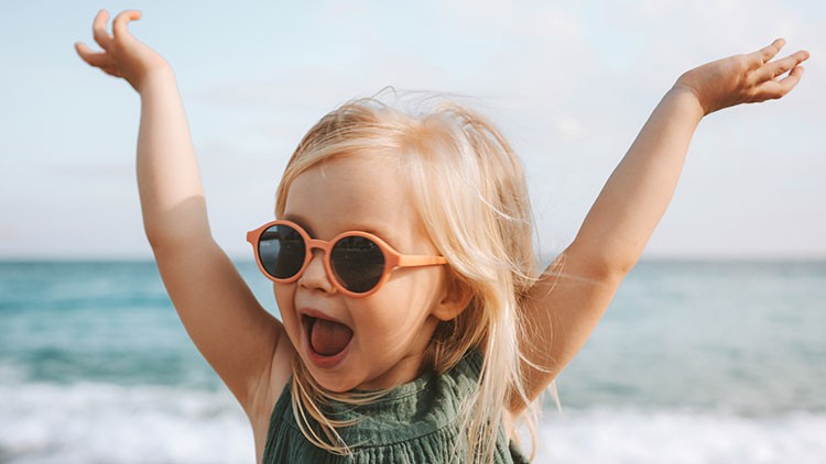 Kind mit langen blonden Haaren trägt eine Sonnenbrille und hebt die Arme vor einem Strand mit Meer und blauem Himmel.