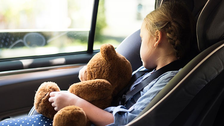 Kind sitzt in einem Kindersitz im Auto und hält einen braunen Teddybären, ein Fenster zeigt unscharfe Außenansicht.