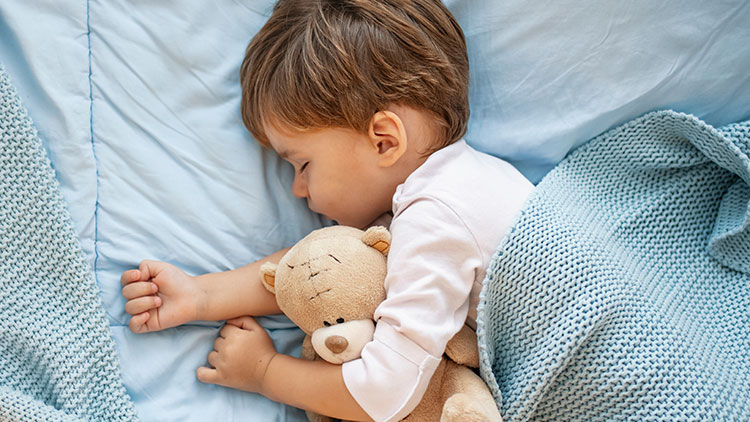 Ein Kind in weißem Oberteil liegt in blauer Bettwäsche mit gestrickter Decke und hält einen braunen Teddybär im Arm.
