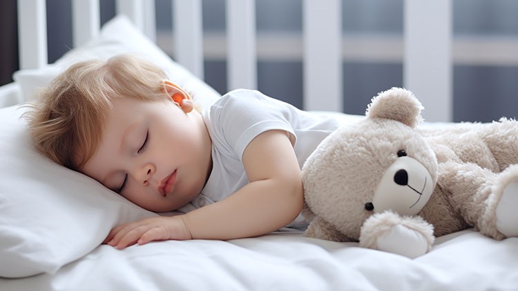 Kind in weißem Schlafanzug liegt auf einem weißen Bett, rechts daneben ein beiger Stoffteddy.