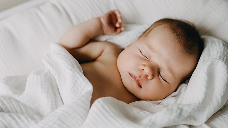 Ein Baby liegt auf einem weißen Stofftuch und ist teils mit einer weißen Decke zugedeckt.
