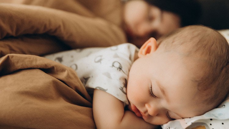 Ein schlafendes Baby in einem hellen Strampler liegt auf einem braunen Bett neben einer Person unter einer braunen Decke.