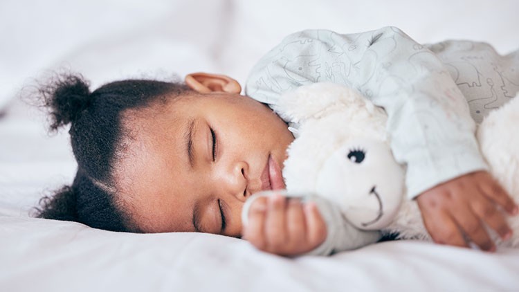 Kind liegt in heller Kleidung auf einem weißen Bett und hält ein weißes Kuscheltier mit aufgesticktem Gesicht in der Hand.