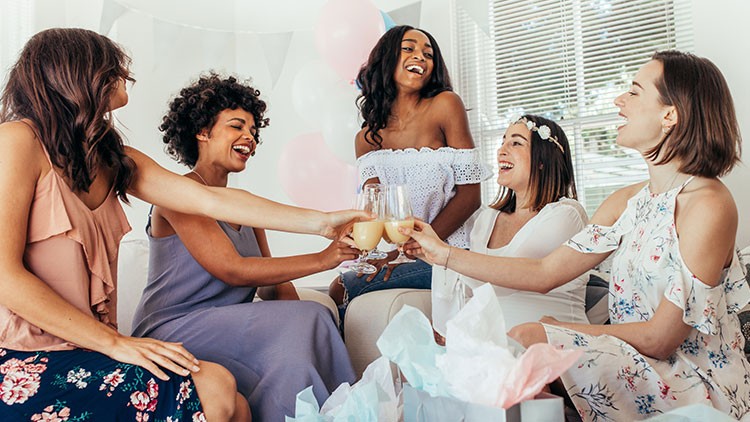 Fünf Personen sitzen zusammen auf einem Sofa, stoßen mit Gläsern an und sind von Luftballons und Geschenken umgeben.