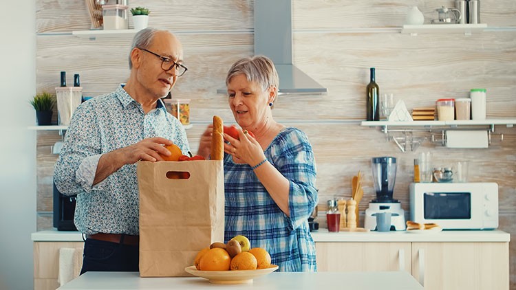Personen entnehmen Lebensmittel wie Brot und Obst aus einer Papiertüte, eine moderne Küche mit Obstschale ist im Hintergrund.