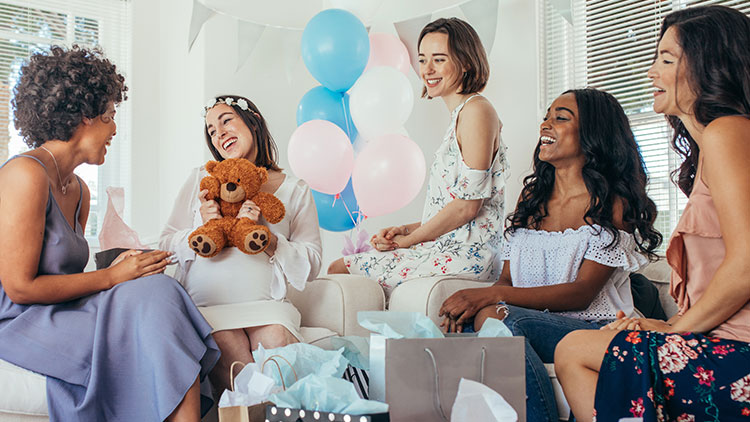 Person hält braunen Teddy in Händen, umgeben von rosa-blauen Luftballons und weiteren sitzenden Personen im Innenraum.