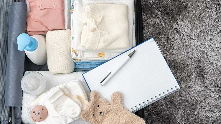 Ein offener Koffer mit Babykleidung, Windeln, einer Flasche, einem Kuscheltier, einem Notizblock und einem Stift liegt auf grauem Teppich.