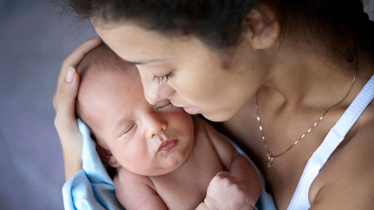 Eine Person hält ein Baby in blauer Decke auf dem Arm, der Hintergrund ist unscharf und dunkelviolett.