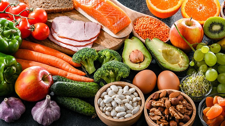 Verschiedene Lebensmittel wie Obst, Gemüse, Lachs, Eier, Hülsenfrüchte, Nüsse, Brot und Schinken auf einem Tisch.