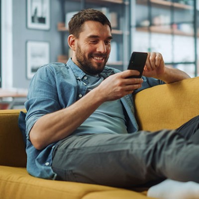 Person sitzt entspannt auf einem gelben Sofa in einem modernen Wohnzimmer und schaut auf ein Smartphone.