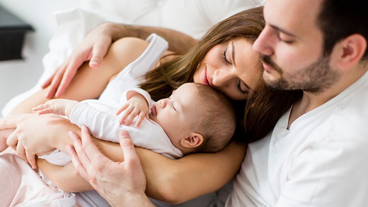 Person in weißem Oberteil hält ein Baby im Arm, während ein weiterer Arm die Person umarmt, weiße Bettwäsche im Hintergrund.
