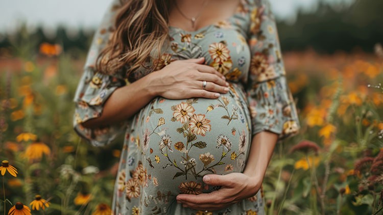 Hände halten einen Schwangerschaftsbauch, der von einem geblümten Kleid bedeckt ist, im Hintergrund Blumenwiese.