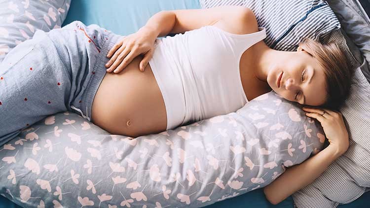 Person in hellem Top liegt seitlich mit einem Schwangerschaftskissen auf gemustertem Bett.