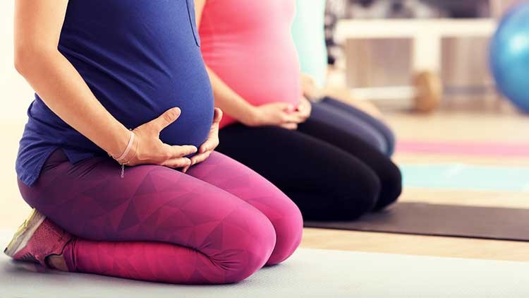 Zwei Personen knien auf Yogamatten, tragen Sportkleidung und halten ihre Hände auf ihre sichtbaren Babybäuche.