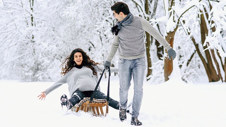 Eine Person sitzt lachend auf einem Holzschlitten im Schnee, eine andere Person zieht den Schlitten durch eine schneebedeckte Landschaft.