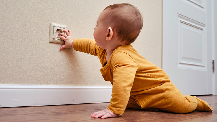 Ein Kind in gelbem Strampler krabbelt auf Holzboden und greift nach einer Steckdose an der Wand.
