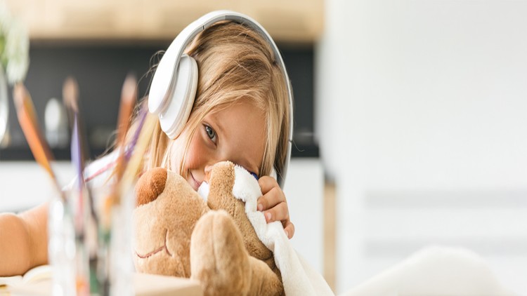 Eine Person trägt weiße Kopfhörer und hält ein braunes Stofftier auf einem Tisch, im Hintergrund ist eine Küche erkennbar.