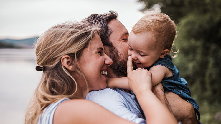 Person hält ein Kind im Arm, im Hintergrund sind Bäume und ein See zu sehen.