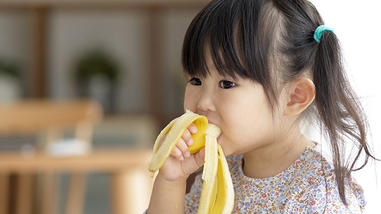 Kind mit Zopf hält eine geschälte Banane in der Hand, im Hintergrund ein verschwommener Innenraum mit Möbeln und Pflanzen.