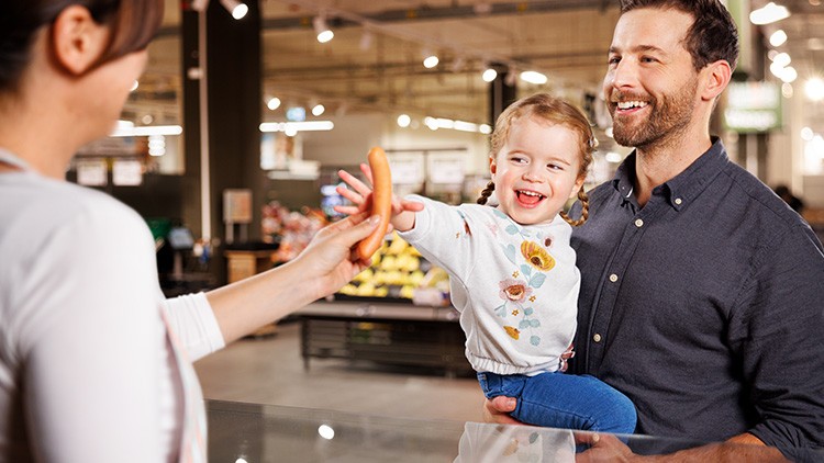 Eine Person reicht einem kleinen Kind eine Wurst, im Hintergrund sind Verkaufsstände mit Obst und Gemüse in einem Markt zu sehen.