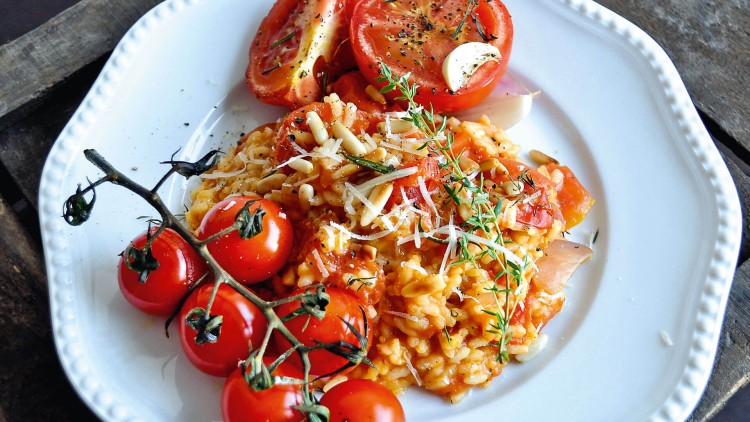Ein weißer Teller mit Tomatenrisotto, frischen Cocktailtomaten am Zweig, geriebenem Käse, Pinienkernen und gebackenen Tomaten.