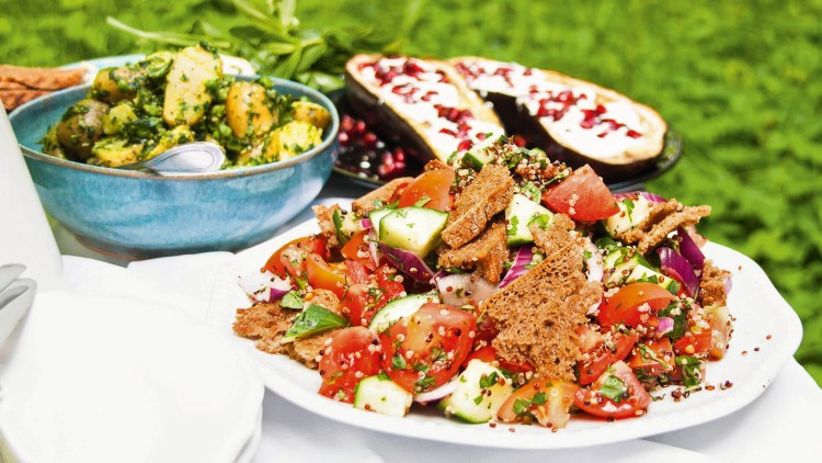 Ein Teller mit buntem Salat aus Tomaten, Gurken, Kräutern und Brotchips, daneben Kräuterkartoffeln und gefüllte Auberginen.