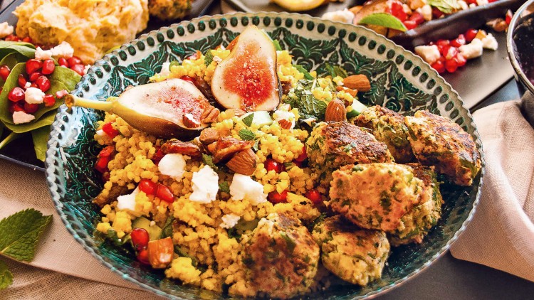 Bunter Couscous-Salat mit Feigen, Granatapfelkernen, Mandeln, Kräutern, Käse und Falafelbällchen auf einem grünen Teller.