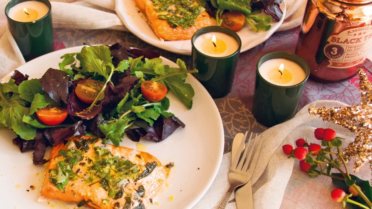 Zwei Teller mit Lachsfilet, grünem Salat und Cherrytomaten, umgeben von Kerzen, Dekoration und Besteck auf einer Tischdecke.