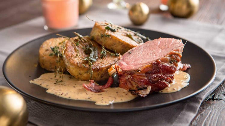 Ein dunkler Teller mit Fleischscheibe, zwei Kartoffelscheiben, körniger heller Sauce, Kräutern, Kerzen und goldenen Kugeln.