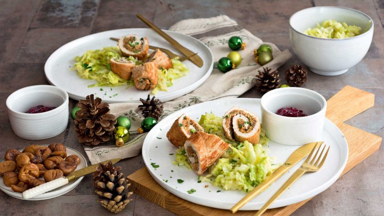 Gefüllte Fleischröllchen mit Kräutern, serviert auf Wirsing, daneben Preiselbeersoße und Maronen, dekoriert mit Tannenzapfen und Kugeln.