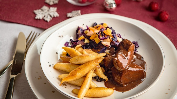 Ein weißer Teller mit Schupfnudeln, Rotkohl mit Apfelstücken und Fleisch in dunkler Sauce auf rot gedecktem Tisch.