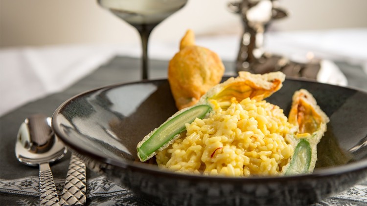 Safran-Risotto in einem schwarzen Teller, dekoriert mit gefüllten Zucchiniblüten und einem Glas Weißwein im Hintergrund.