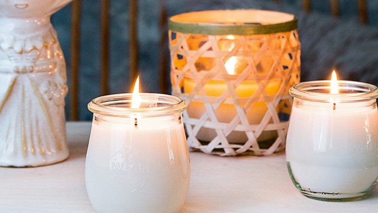 Drei weiße Kerzen in Gläsern und ein Kerzenhalter neben einer weißen Vase mit Eukalyptuszweigen auf hellem Tisch.
