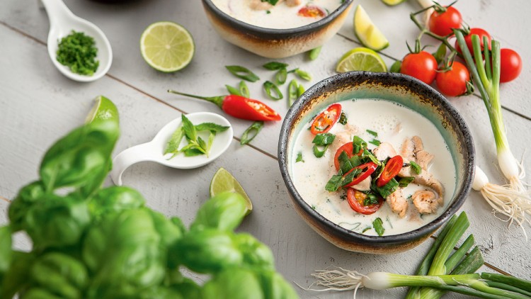 Zwei Schalen mit Kokossuppe, verziert mit Chili, Tomate und Kräutern, umgeben von Limetten, Frühlingszwiebeln und Basilikum.
