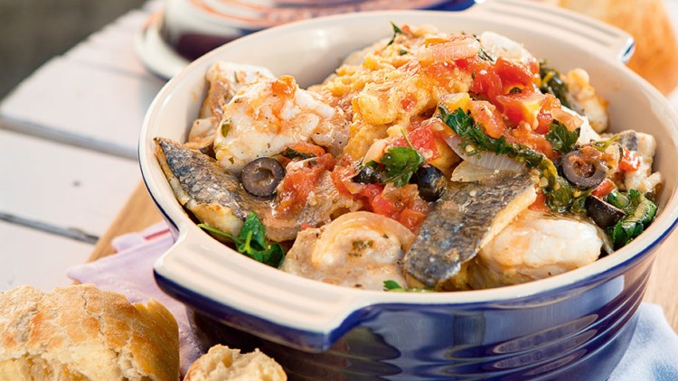 Auf einem weißen Tisch steht eine blaue Auflaufform mit Fisch, Gemüse und Tomatensoße, daneben Brot und ein Salzschälchen.