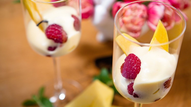 Zwei Dessertgläser mit heller Creme, Himbeeren und gelben Fruchtstücken auf Holztisch, im Hintergrund rosa Blumen.