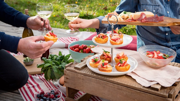 Picknick mit Häppchen auf einer Etagere, Erdbeeren, Blaubeeren, Basilikum und zwei Personen mit Weißweingläsern.