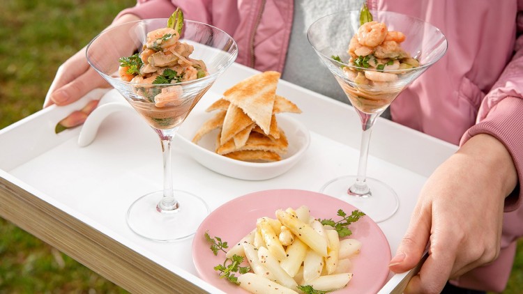 Eine Person trägt ein Tablett mit zwei Gläsern Garnelencocktail, geröstetem Toast und weißem Spargel auf einem rosa Teller.